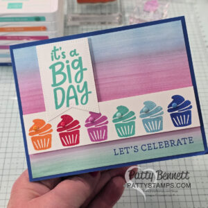 A hand holds a colorful greeting card with a watercolor background in blue, purple, and pink. The card features six rainbow cupcakes, the phrases "it's a Big DAY" and "LET'S CELEBRATE," and the website PattyStamps.com. Craft supplies are seen behind. To read more, visit Patty Stamps (pattystamps.com)