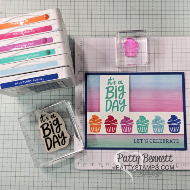 A handmade card with “It’s a Big Day” and colorful cupcake stamps is displayed on a grid mat. Ink pads in various colors are stacked on the left. Two clear acrylic stamps—one with the cupcake design, one with the card’s main text—are next to the card. To read more, visit Patty Stamps (pattystamps.com)