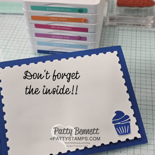 A blue-bordered card with a white center reads "Don't forget the inside!!" in black script. There’s a small blue cupcake stamp in the lower right corner. In the background, stacked colorful ink pads are visible on a grid-patterned craft table. To read more, visit Patty Stamps (pattystamps.com)