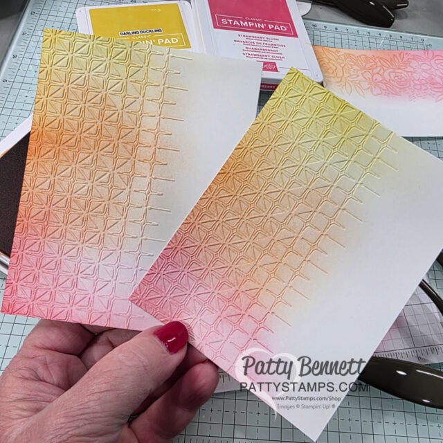 A hand holds two embossed paper cards with the Glass & Gardens Embossing Folder geometric star pattern, both inked in gradient shades of pink, orange, and yellow. Ink pads, scissors, and crafting supplies are visible on the work surface. The watermark reads "Patty Bennett PattyStamps.com. To read more, visit Patty Stamps (pattystamps.com)