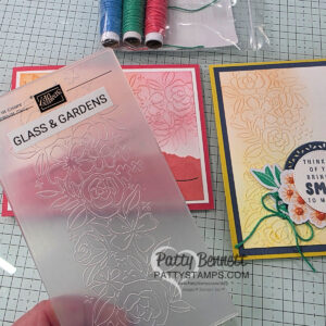 A hand holds a floral-patterned embossing folder labeled "Glass & Gardens" over a gridded work surface. Nearby are two handmade greeting cards with floral embossing, colored twine, and a bag with spools of red, green, and blue string. "PattyStamps.com" watermark is visible. To read more, visit Patty Stamps (pattystamps.com)