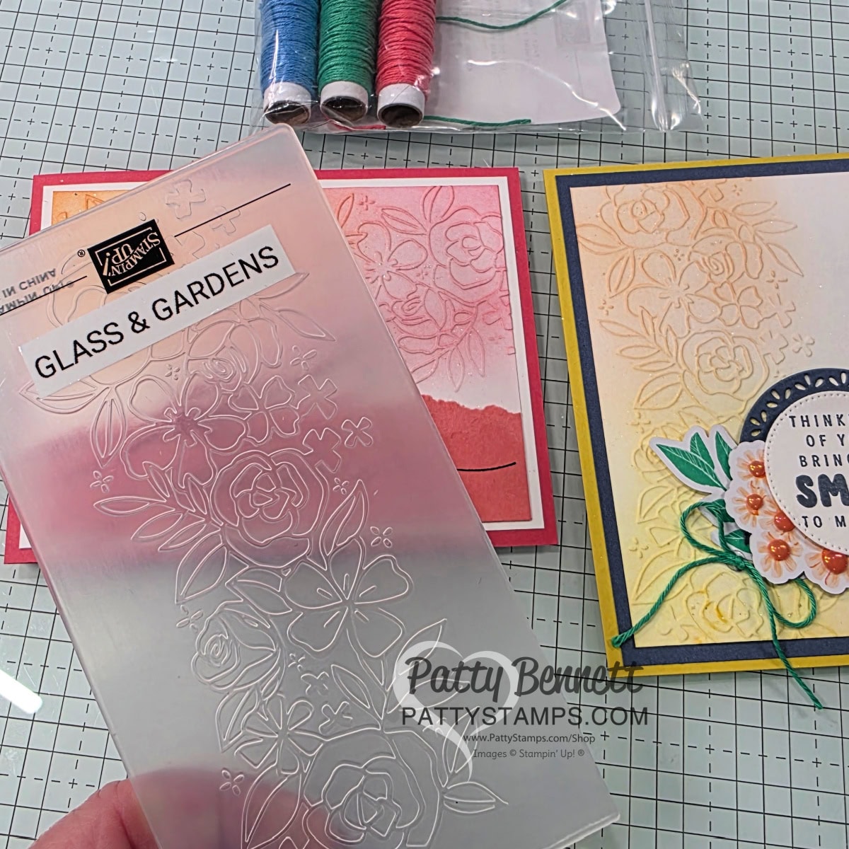A hand holds a floral-patterned embossing folder labeled "Glass & Gardens" over a gridded work surface. Nearby are two handmade greeting cards with floral embossing, colored twine, and a bag with spools of red, green, and blue string. "PattyStamps.com" watermark is visible. To read more, visit Patty Stamps (pattystamps.com)