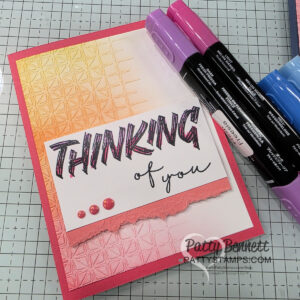 A handmade card with a textured, gradient background in yellow, orange, and pink tones. Bold text reads "THINKING" above script "of you" on a white panel with a torn edge and three pink dots. Purple and pink markers are nearby. To read more, visit Patty Stamps (pattystamps.com)
