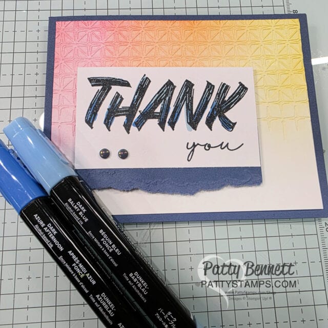 A handmade thank you card with bold, blue "THANK" and cursive "you" on textured white paper, with blue sequins and a torn blue edge. The background fades from yellow to pink. Three blue markers lie in front. Grid-lined craft mat underneath. To read more, visit Patty Stamps (pattystamps.com)