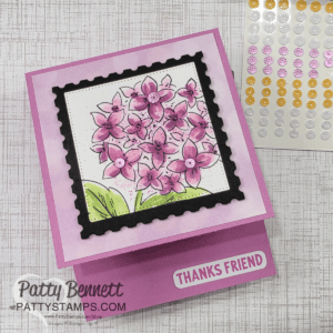 A handmade purple card featuring a floral design. The card showcases pink flowers within a black, postage-stamp-style frame on the front. "THANKS FRIEND" is printed on a raised section. A sheet of colorful adhesive gems is nearby. The card is signed by Patty Bennett from PattyStamps.com. To read more, visit Patty Stamps (pattystamps.com)