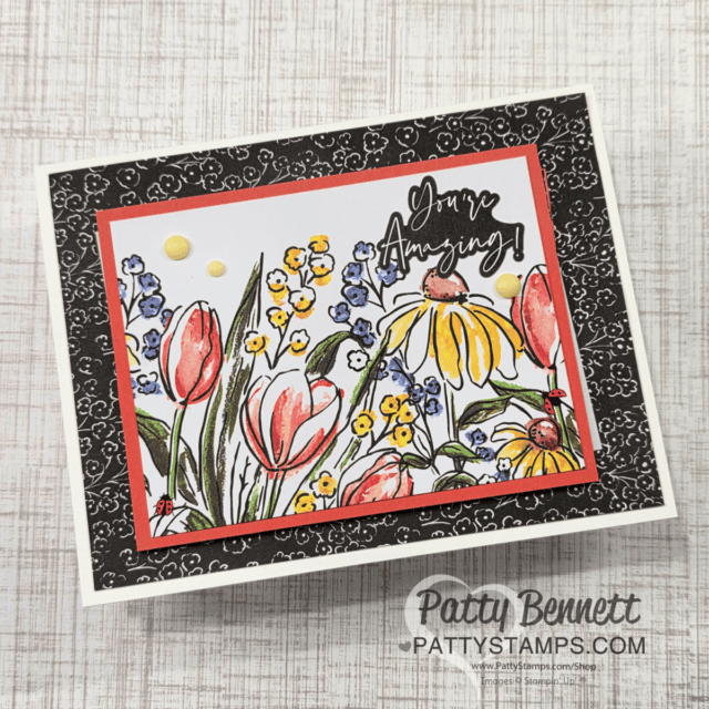 A handmade card with a floral design bordered by a black and white floral pattern. The center artwork depicts red tulips, yellow daisies, and blue flowers. The text "You're Amazing" is written in black cursive. The card is signed by Patty Bennett from Patty Stamps. To read more, visit Patty Stamps (pattystamps.com)