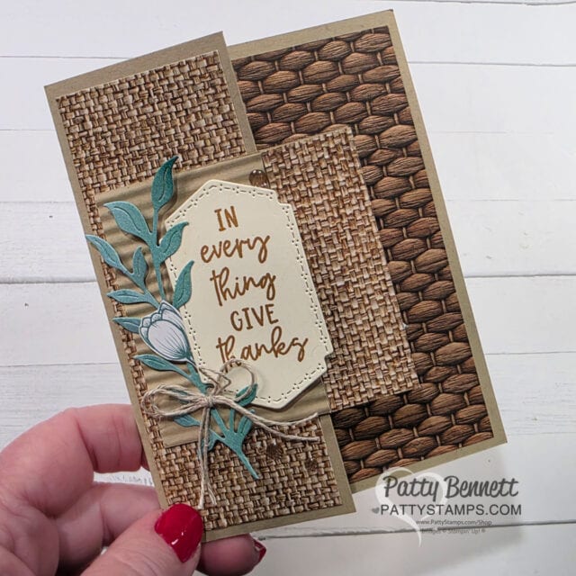 A hand holds a handmade card with woven-texture patterns in tan and brown. A tag with gold script reads "IN every thing GIVE thanks," decorated with green leaves, a white flower, and a twine bow. "Patty Bennett PATTYSTAMPS.com" is visible at the bottom. To read more, visit Patty Stamps (pattystamps.com)