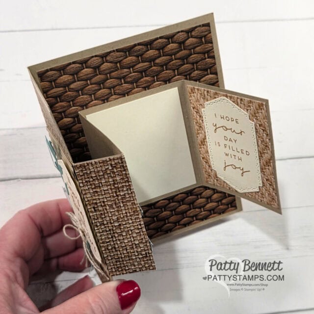 A hand holds an intricate, handmade card with woven textures and neutral tones. The card is open, revealing a message on a tag that says, "I hope your day is filled with joy," and a blank note section inside. The background is a light wood table. To read more, visit Patty Stamps (pattystamps.com)