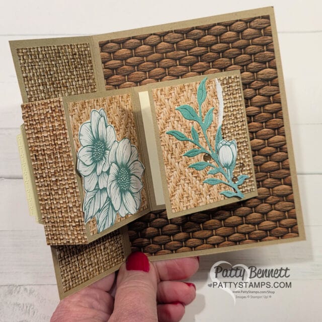 A hand holds a layered handmade card with woven basket patterns. The card features two square panels: one with sketched blue flowers, and another with leafy blue-green die-cuts. The background is brown, textured, and the card is photographed on a white surface. To read more, visit Patty Stamps (pattystamps.com)