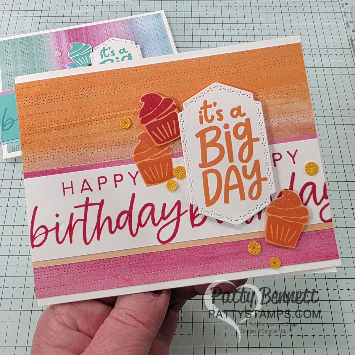 Two handmade birthday cards, each decorated with a “It’s a Big Day” greeting from Stampin UP! Around it All and With You in Mind stamp sets, striped backgrounds in teal or orange tones, cupcake cutouts, and sequins. A package of starburst sequins in yellow, pink, and white sits beside the cards on a grid-patterned surface. To read more, visit Patty Stamps (pattystamps.com)