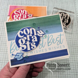 A hand holds a handmade card featuring a blue and white "congrats" circle from Stampin' Up! Around it All bundle, a small graduation cap, and blue stripes with "Best Wishes Wishes Wishes" in the background. Two more similar cards and a stamp are on a textured, gridded workspace. To read more, visit Patty Stamps (pattystamps.com)