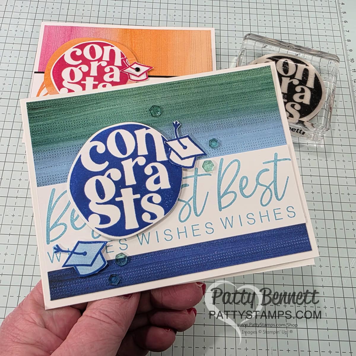 A hand holds a handmade card featuring a blue and white "congrats" circle from Stampin' Up! Around it All bundle, a small graduation cap, and blue stripes with "Best Wishes Wishes Wishes" in the background. Two more similar cards and a stamp are on a textured, gridded workspace. To read more, visit Patty Stamps (pattystamps.com)