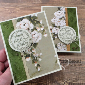 Two handmade birthday cards with floral designs are shown. Both feature painted white flowers on a green and tan background, a “Happy Birthday Wishes” circular label, and a twine bow. The cards have a textured, elegant look and rest on a wood surface. To read more, visit Patty Stamps (pattystamps.com)