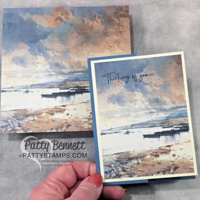 Design #5 of 4 cards with 6x6 DSP. A hand holds a greeting card with a painted seascape and the words "Thinking of you..." The card matches the background paper, which also features a tranquil, pastel-toned shoreline with boats, cloudy sky, and soft, muted colors. "Patty Bennett" watermark appears below. To read more, visit Patty Stamps (pattystamps.com)
