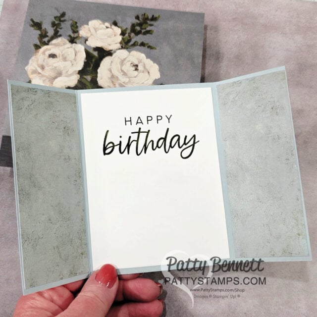 A hand holds a tri-fold birthday card. The card has soft gray textured panels on each side and a white center panel with “HAPPY birthday” written in black script font. In the background, there’s an image of white roses. The watermark reads “Patty Bennett PattyStamps.com.”. To read more, visit Patty Stamps (pattystamps.com)