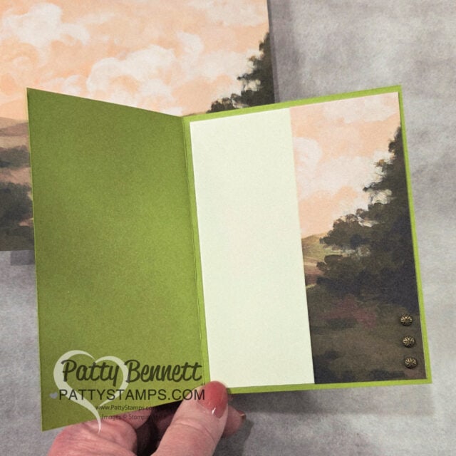 A hand holds open a green card with an inner panel featuring a landscape illustration of trees, hills, and a cloudy sky. The left side has a blank cream section for writing. Three small gold embellishments are aligned vertically in the bottom right corner. To read more, visit Patty Stamps (pattystamps.com)