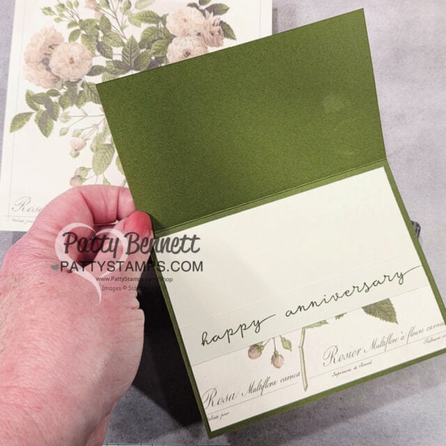A hand holds open a green greeting card with a cream insert. The inside features elegant script reading "happy anniversary" and a botanical design strip. In the background is a floral card with cream roses and green leaves. "Patty Bennett PATYSTAMPS.COM" watermark overlays the image. To read more, visit Patty Stamps (pattystamps.com)