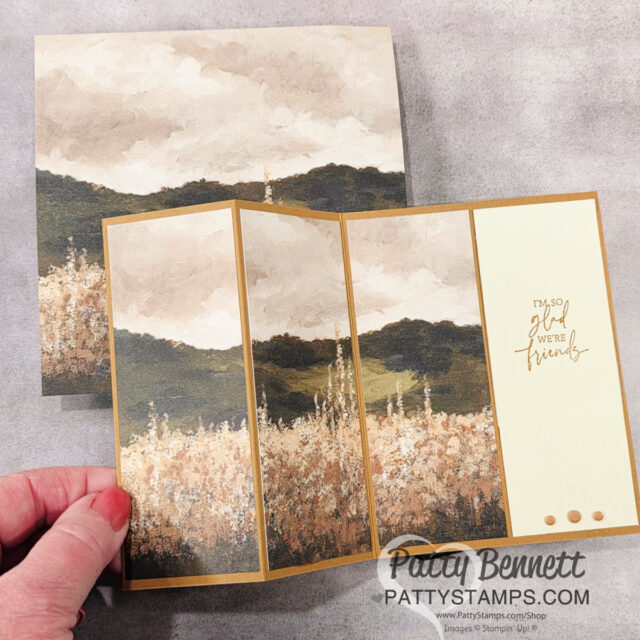 A hand holds a handmade fold-out greeting card featuring a painted landscape of hills and wildflowers in muted earth tones. One panel reads "I'm so glad we're friends" in gold script. The matching envelope lies underneath. Logo text: Patty Bennett, PattyStamps.com. To read more, visit Patty Stamps (pattystamps.com)