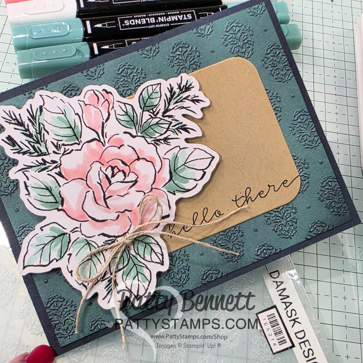 A handmade greeting card features a textured teal background, a large watercolor-style pink rose with green leaves, and a tan card with “Hello There” written in script. The card is embellished with twine and surrounded by markers and craft supplies. To read more, visit Patty Stamps (pattystamps.com)
