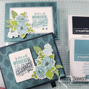 Two handmade greeting cards with blue-green floral designs and layered paper elements. Both cards display the message, "It's a wonderful day to celebrate you," in teal text. Ink pads and crafting tools are visible beside the cards on a gridded work surface. To read more, visit Patty Stamps (pattystamps.com)