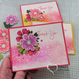 A hand holds a handmade card with pink and red layered flowers and green leaves. The card features embossed and ink-blended floral designs and the words "Thank You" in pink script. Two similar cards with yellow and pink borders are placed on a grid work surface. To read more, visit Patty Stamps (pattystamps.com)