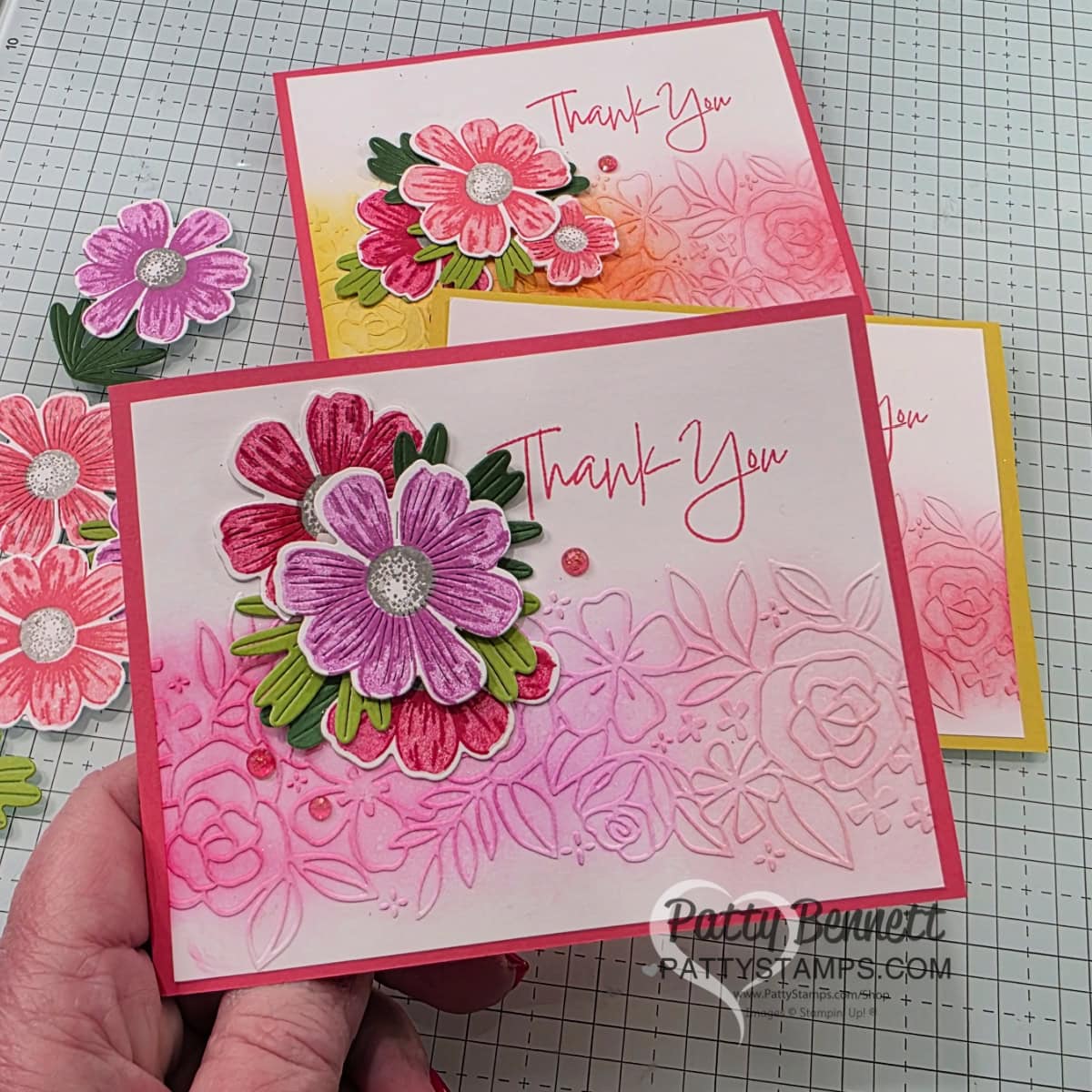 A hand holds a handmade card with pink and red layered flowers and green leaves. The card features embossed and ink-blended floral designs and the words "Thank You" in pink script. Two similar cards with yellow and pink borders are placed on a grid work surface. To read more, visit Patty Stamps (pattystamps.com)