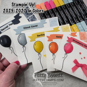 A hand holds four cards displaying Stampin’ Up! 2025-2027 In Colors: Secret Sea, Cloud Cover, Darling Daffodil, Timid Tangerine, and Strawberry Slush. Each card features a balloon and color swatch. Color markers and bows are shown above. To read more, visit Patty Stamps (pattystamps.com)