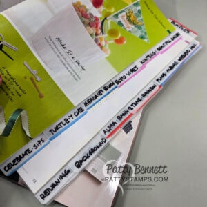 A stack of magazines is shown from the side, each with a handwritten white tab labeling sections such as “CELEBRATE SIP,” “TURTLEY CUTE MEMORIES,” “ALPHA,” “KRAFTY,” and other crafting themes. A “PattyStamps.com” watermark is visible. To read more, visit Patty Stamps (pattystamps.com)