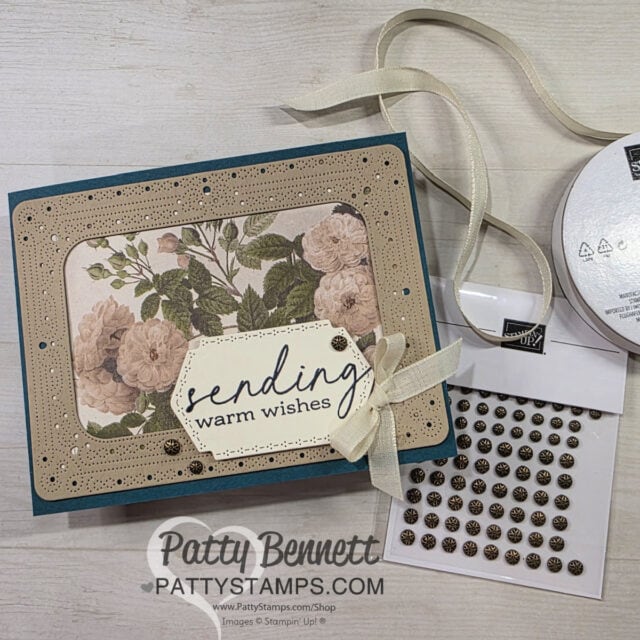 A handmade greeting card decorated with cream and tan floral paper, ornate beige borders, and a white ribbon bow. The card says "sending warm wishes" in script, accented with bronze embellishments. Craft supplies are nearby, including pearl stickers and ribbon. To read more, visit Patty Stamps (pattystamps.com)