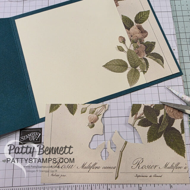 A handmade card in progress lies on a grid workspace. A cream insert with vintage-style rose and leaf illustrations is partially inside a dark green card. Below, cutout floral paper scraps and craft tools are visible. The workspace has Patty Bennett branding. To read more, visit Patty Stamps (pattystamps.com)