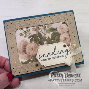 A hand holds a handmade card with a floral background, beige perforated frame, and a cream tag reading "sending warm wishes" in script. The tag is accented with small gold embellishments and a cream ribbon. "Patty Bennett" branding is on the lower right. To read more, visit Patty Stamps (pattystamps.com)