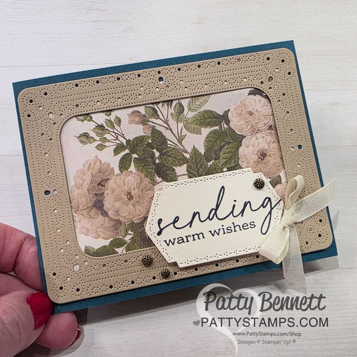 A hand holds a handmade card with a floral background, beige perforated frame, and a cream tag reading "sending warm wishes" in script. The tag is accented with small gold embellishments and a cream ribbon. "Patty Bennett" branding is on the lower right. To read more, visit Patty Stamps (pattystamps.com)