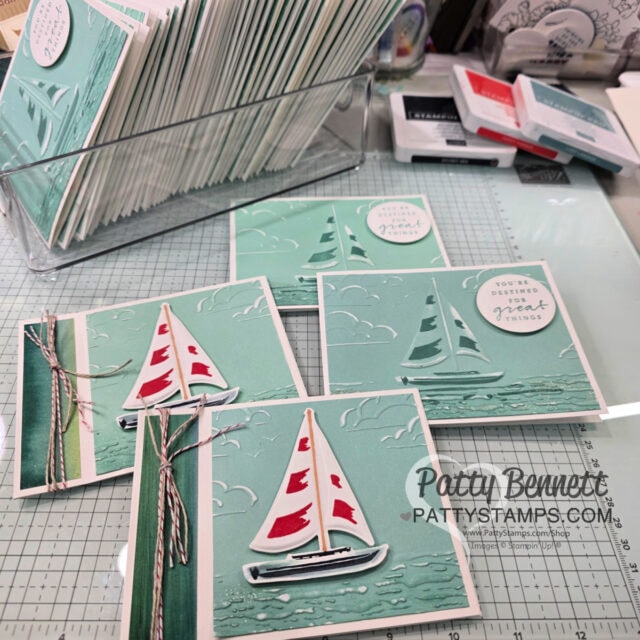 Handmade greeting cards with embossed teal backgrounds and raised sailboat designs, some with red-sailed boats. One card is tied with twine. Stamped sentiments are visible. Numerous cards are stacked in a clear tray; crafting supplies and ink pads are nearby. To read more, visit Patty Stamps (pattystamps.com)