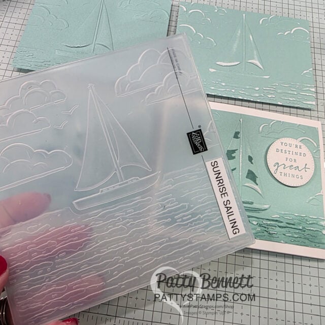 A hand holds a clear "Sunrise Sailing" embossing folder with a sailboat and clouds design. In the background are several light blue embossed sailing-themed cards, one reading "You're destined for great things." The surface has a grid and "Patty Bennett" watermark. To read more, visit Patty Stamps (pattystamps.com)