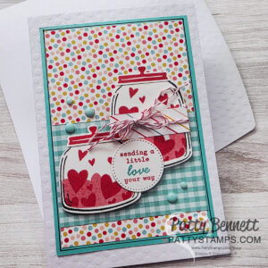 A colorful handmade card with two jars decorated with red hearts, a round tag reading “sending a little love your way,” baker’s twine, and pastel blue dots. The background is polka-dotted and checkered, with the card set on a white envelope. To read more, visit Patty Stamps (pattystamps.com)