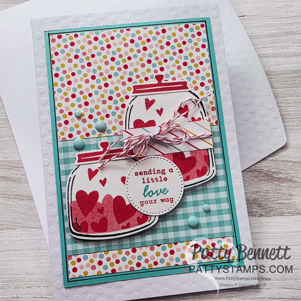 A colorful handmade card with two jars decorated with red hearts, a round tag reading “sending a little love your way,” baker’s twine, and pastel blue dots. The background is polka-dotted and checkered, with the card set on a white envelope. To read more, visit Patty Stamps (pattystamps.com)