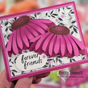 A handmade greeting card with two large, layered pink and purple paper coneflowers on a black-and-white leafy background. The card includes the handwritten text "forever friends." It is signed "Patty Bennett PATTYSTAMPS.com" at the bottom right. To read more, visit Patty Stamps (pattystamps.com)
