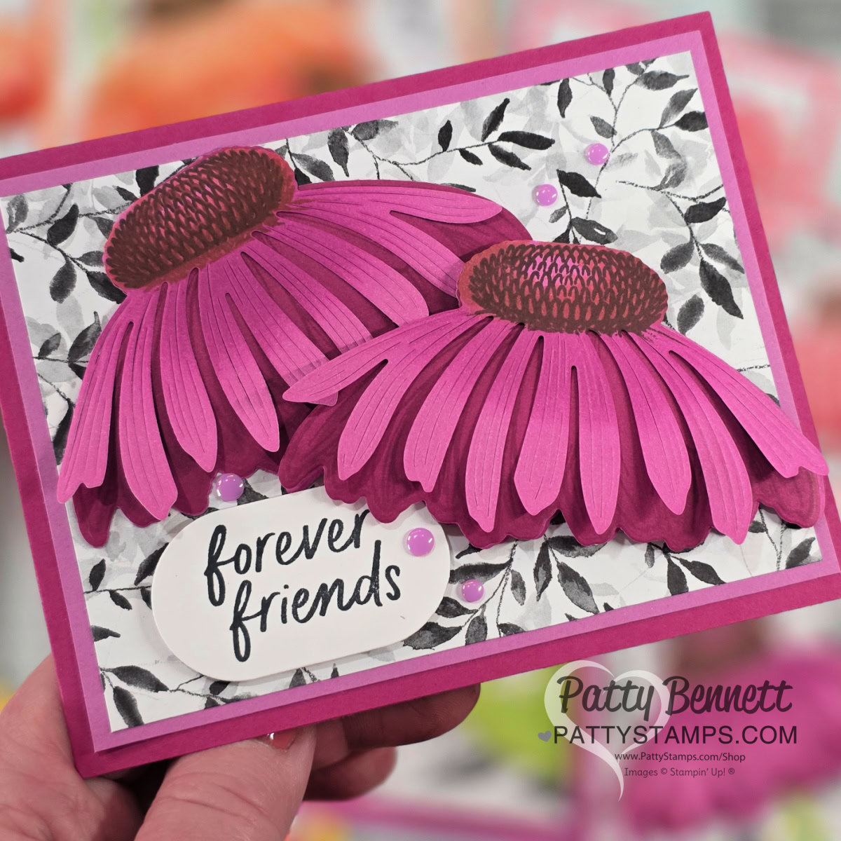 A handmade greeting card with two large, layered pink and purple paper coneflowers on a black-and-white leafy background. The card includes the handwritten text "forever friends." It is signed "Patty Bennett PATTYSTAMPS.com" at the bottom right. To read more, visit Patty Stamps (pattystamps.com)