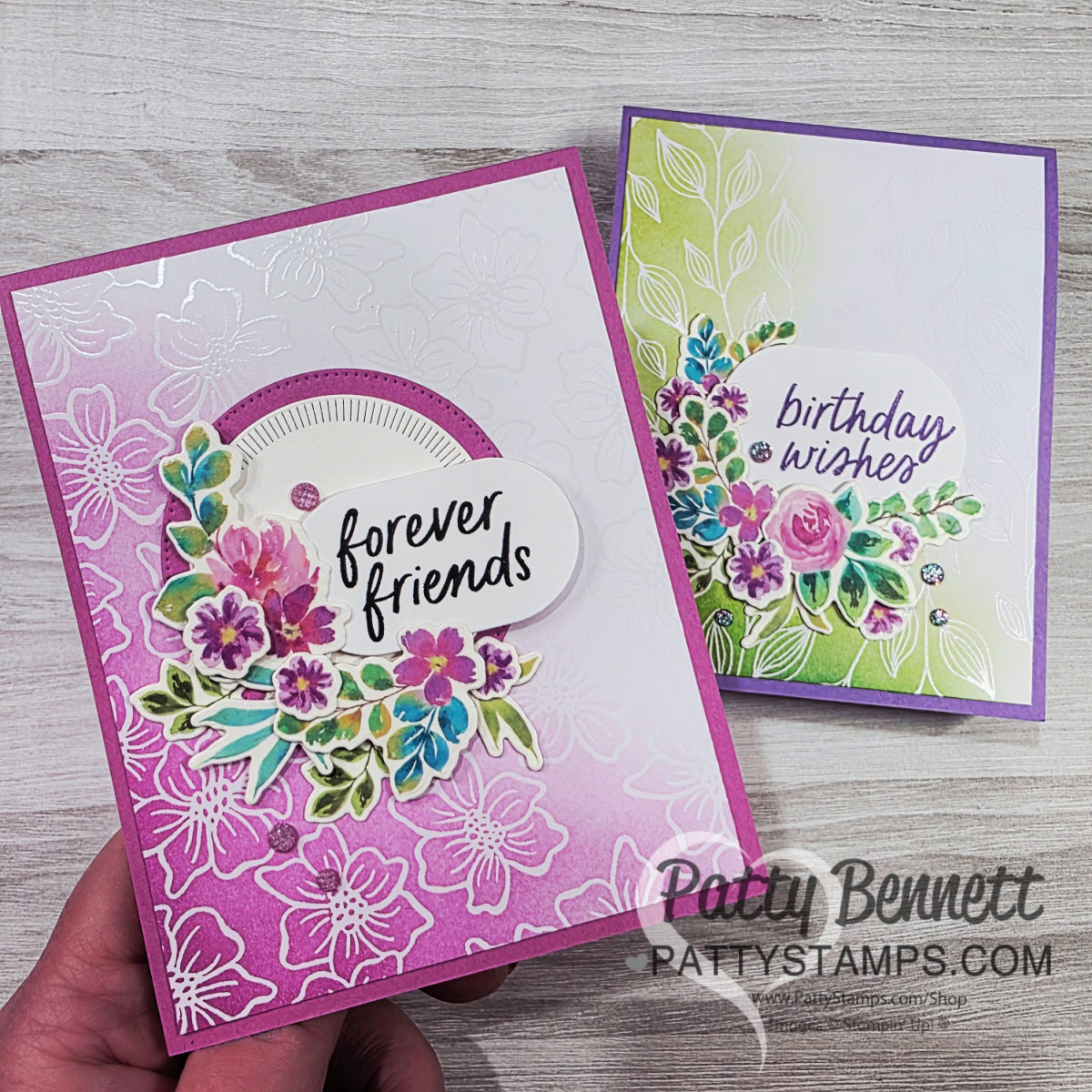 Two handmade greeting cards with floral designs. The left card is pink and white with “forever friends” and layered flowers. The right card is green and white with “birthday wishes” and purple flowers. A hand holds both cards on a light wooden surface. To read more, visit Patty Stamps (pattystamps.com)