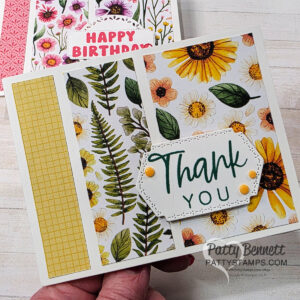 A handmade card with “Thank You” in green text on a white tag, decorated with yellow and orange flowers and leaves. The left side features a yellow grid pattern strip. Another floral "Happy Birthday" card is partially visible in the background. To read more, visit Patty Stamps (pattystamps.com)