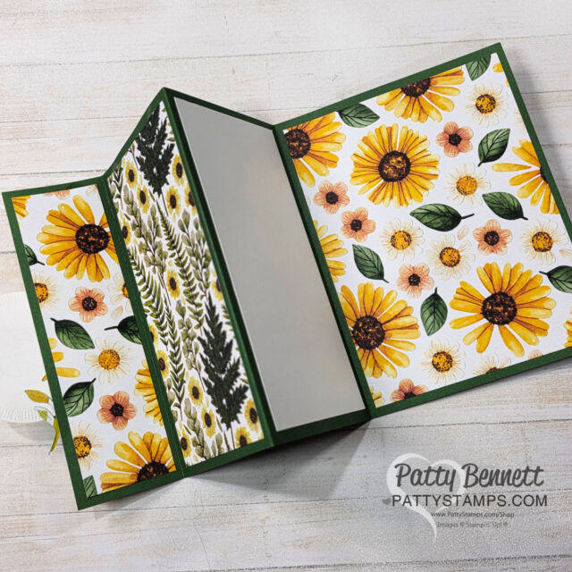 A handmade card with fold-out panels is displayed open on a white surface. The card features yellow sunflower and daisy patterns, green leaves, and a central blank panel for writing. The website "PattyStamps.com" and "Patty Bennett" are printed in the lower right corner. To read more, visit Patty Stamps (pattystamps.com)
