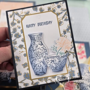 A hand holds an ornate birthday card featuring a blue and white vase and bowl with floral patterns, and an orange flower. The card has "HAPPY BIRTHDAY" in gray, a gold-bordered frame, and a background of blue and gold flowers. To read more, visit Patty Stamps (pattystamps.com)