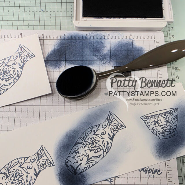 A crafting workspace with a grid mat, ink pad, blending brush, and stamped vases on white paper. Blue ink is blended around the vase images, and the website “PattyStamps.com” is printed on the paper. Some ink smudges are visible on the mat. To read more, visit Patty Stamps (pattystamps.com)
