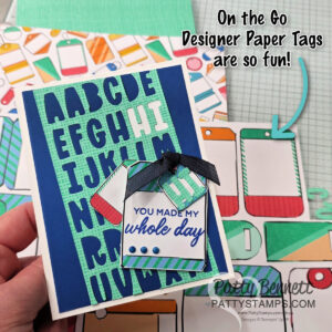A hand holds a handmade card featuring large blue alphabet cutouts on a green grid, with layered phone-shaped tags reading "You made my whole day." Colorful phone designs and craft supplies are visible in the background, with playful text pointing to the tags. To read more, visit Patty Stamps (pattystamps.com)