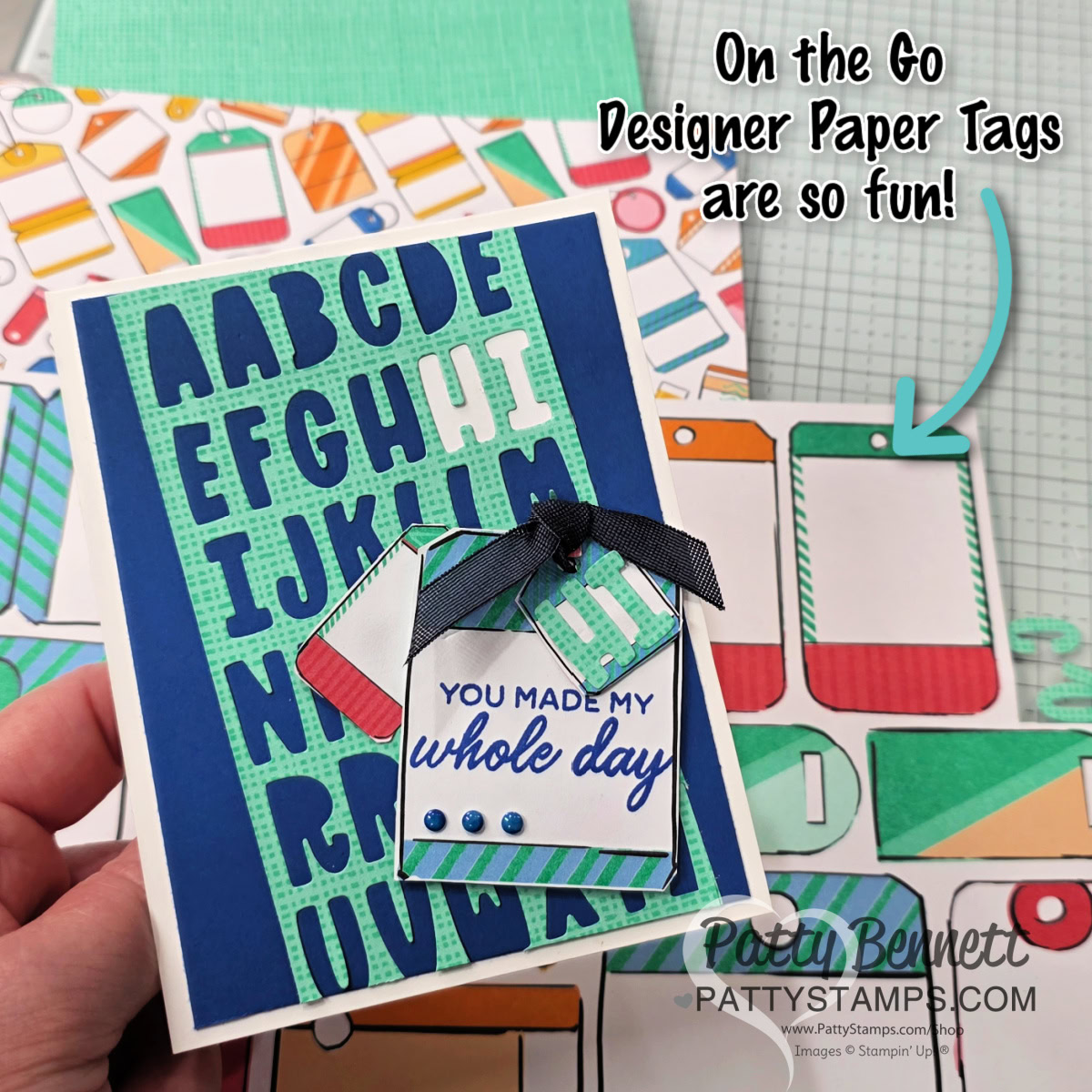 A hand holds a handmade card featuring large blue alphabet cutouts on a green grid, with layered phone-shaped tags reading "You made my whole day." Colorful phone designs and craft supplies are visible in the background, with playful text pointing to the tags. To read more, visit Patty Stamps (pattystamps.com)