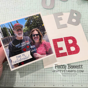Party Alphabet anniversary card. A hand holds a homemade card featuring a photo of an older man and a woman smiling outdoors. The card says "TIME TO celebrate!" Cutout letters "EB" in silver and red are on the card and table. The surface has a cutting mat and the website "PattyStamps.com. To read more, visit Patty Stamps (pattystamps.com)