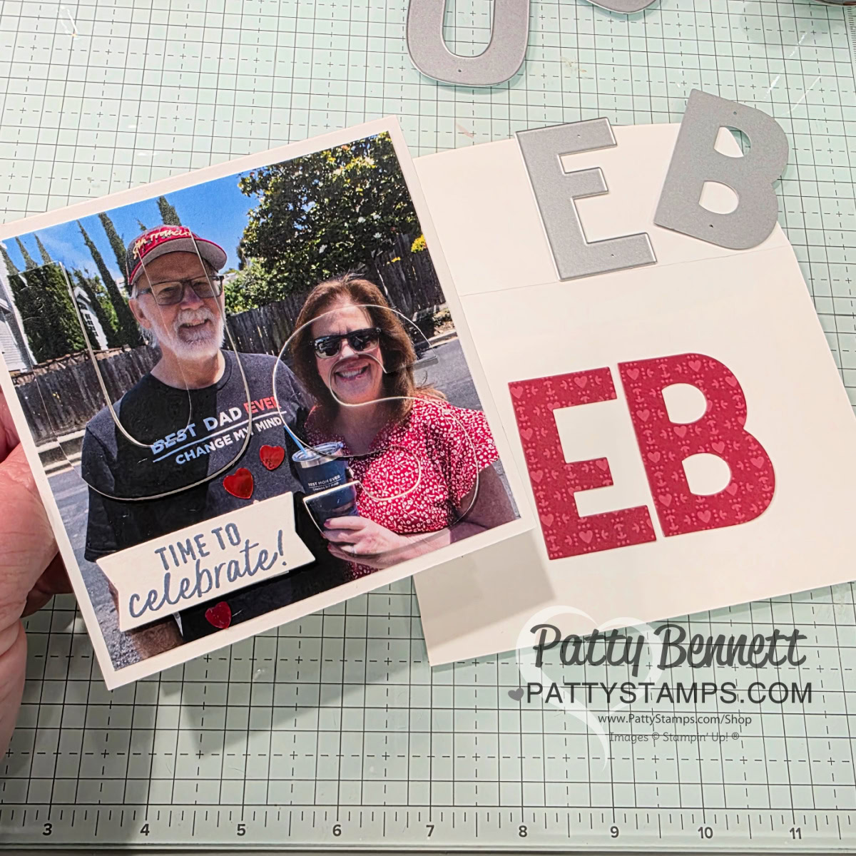 Party Alphabet anniversary card. A hand holds a homemade card featuring a photo of an older man and a woman smiling outdoors. The card says "TIME TO celebrate!" Cutout letters "EB" in silver and red are on the card and table. The surface has a cutting mat and the website "PattyStamps.com. To read more, visit Patty Stamps (pattystamps.com)