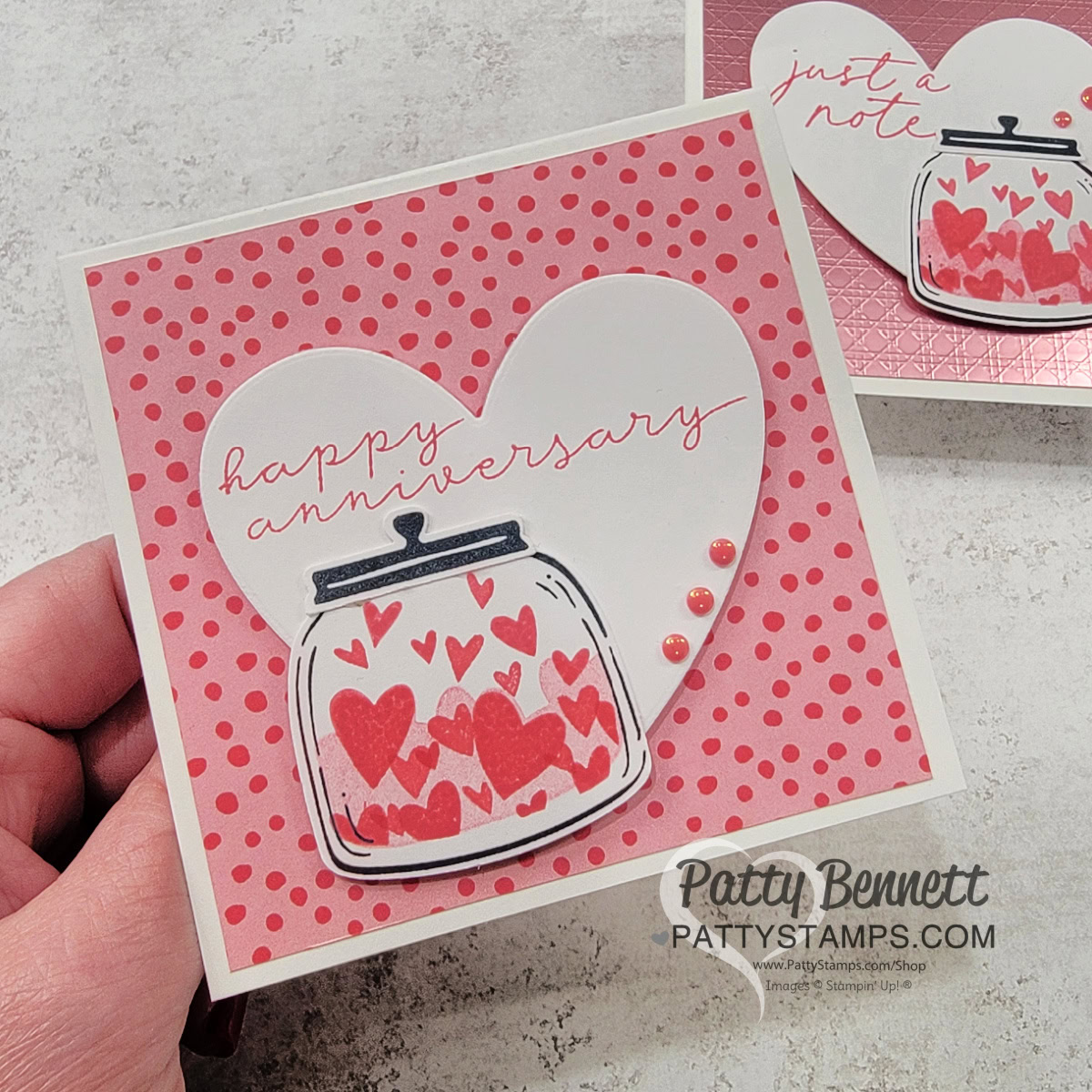 A hand holds a sweet square handmade heart card with a pink polka dot background. The card features a large white heart with "happy anniversary" written in script, and a jar illustration filled with red and pink hearts. Three pink pearls accent the lower right. To read more, visit Patty Stamps (pattystamps.com)