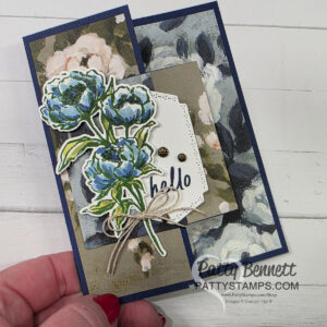 Beautiful Gallery Hidden Door Card A handmade greeting card features layered floral designs with blue and white peonies, a “hello” message on a tag, and a twine bow. The background has dark blue and beige flower patterns. A hand holds the card. The watermark reads “Patty Bennett PATTYSTAMPS.com.”. To read more, visit Patty Stamps (pattystamps.com)
