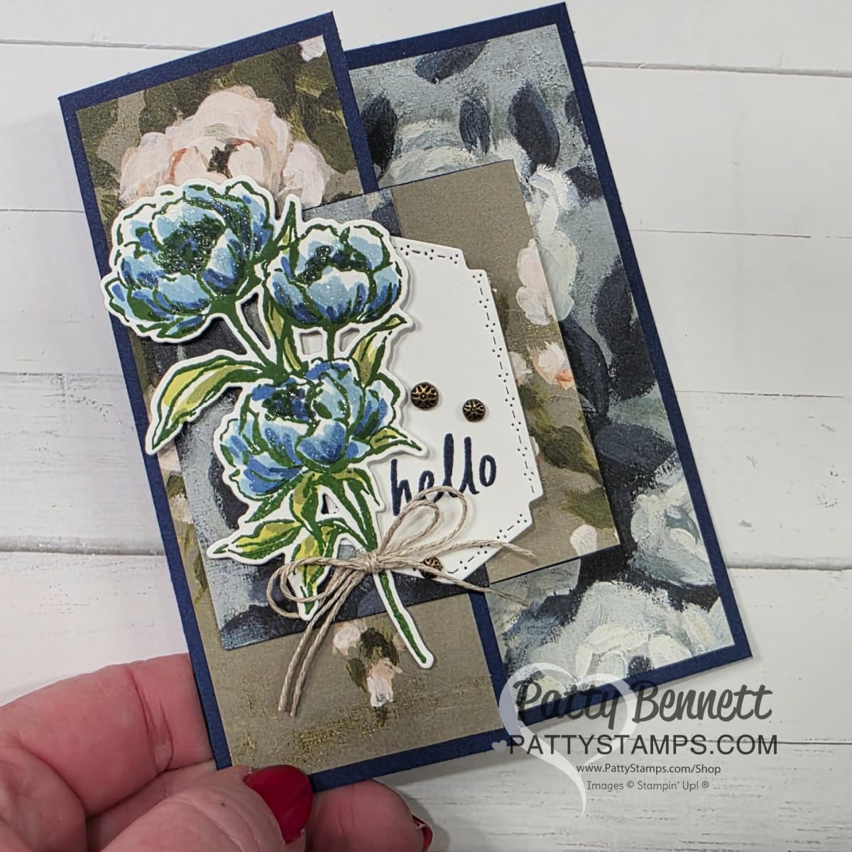 Beautiful Gallery Hidden Door Card A handmade greeting card features layered floral designs with blue and white peonies, a “hello” message on a tag, and a twine bow. The background has dark blue and beige flower patterns. A hand holds the card. The watermark reads “Patty Bennett PATTYSTAMPS.com.”. To read more, visit Patty Stamps (pattystamps.com)